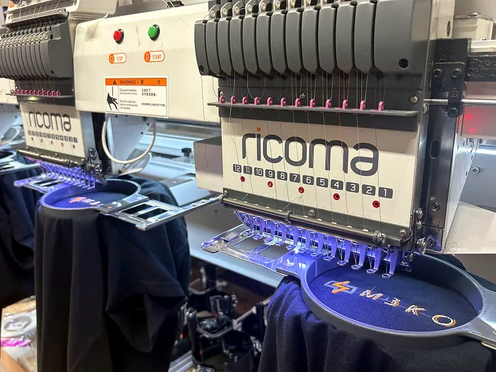 Вышивальные машины Ricoma вышивают логотип на тёмно-синем изделии в пяльцах